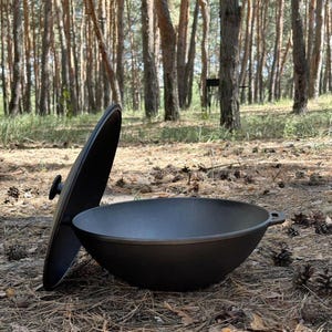 May include: A black cast iron wok with a rounded bottom and a matching black lid leaning against its side. The wok features a small handle on one side and a central knob on the lid, showcasing a smooth, dark, and durable metallic construction.