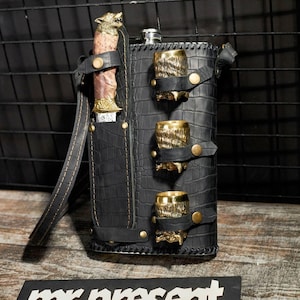 May include: A black leather flask with a textured finish, featuring three gold-coloured shot glasses and a knife with a wolf head handle. The flask has a strap and is secured with gold-coloured studs.