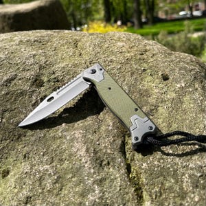 Könnte beinhalten: Ein teilweise geöffnetes Klappmesser mit silberner Klinge und gezahnter Kante. Der Griff ist olivgrün mit strukturiertem Muster und silbernen Akzenten. Eine schwarze Kordel ist am Griff befestigt.