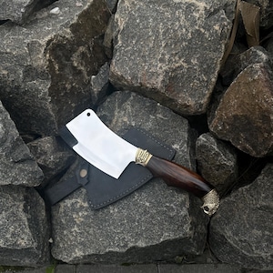 Könnte beinhalten: Ein Edelstahl-Hackbeil mit dunklem Holzgriff und Messingakzenten, auf einer schwarzen Lederscheide. Der Griff hat ein dekoratives Messing-Endstück. Das Hackbeil wird vor dem Hintergrund grauer Felsen präsentiert.