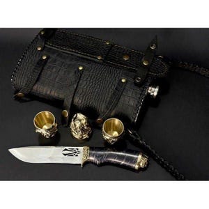 Könnte beinhalten: Eine schwarze Lederflasche mit Krokodiltextur, Messingakzenten und einer passenden Tragetasche. Enthält ein dekoratives Messer mit gemusterter Klinge und zwei kleine goldfarbene Becher. Eine kleine goldfarbene Skulptur ist ebenfalls enthalten.