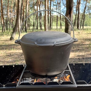 Puede incluir: Una olla holandesa de hierro fundido negro con tapa y asa metálica, colocada sobre una parrilla con brasas ardientes. La olla es redonda y tiene una superficie texturizada, ideal para cocinar al aire libre.