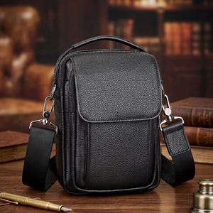Könnte beinhalten: Schwarze Leder-Umhängetasche mit Tragegriff und verstellbarem Schultergurt. Die Tasche hat einen Klappverschluss und eine Vordertasche. Die Beschläge sind silberfarben. Die Tasche ist etwa 23 cm hoch und 18 cm breit.