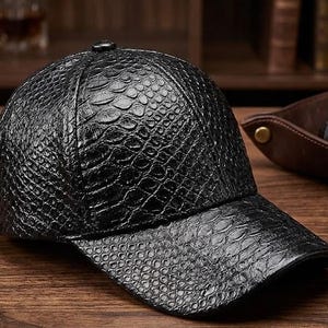 Op de afbeelding: Een zwarte baseballcap met een getextureerd, reptielachtig patroon. De cap heeft een gebogen klep en een kleine knoop aan de bovenkant. De hoed is gemaakt van een donker materiaal en lijkt van leer te zijn. De cap ligt op een houten oppervlak.