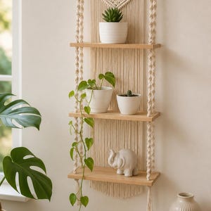 Puede incluir: Estantería de pared de macramé con tres estantes de madera, con plantas en macetas y una pequeña figura de elefante. El macramé es beige, los estantes tienen un acabado de madera natural. Un elemento decorativo y funcional.