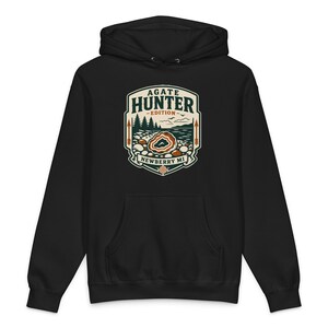 May include: Black hooded sweatshirt featuring a graphic with the text "AGATE HUNTER - EDITION - NEWBERRY MI." The design includes a landscape with trees, a lake, and an agate stone. The sweatshirt has a front pocket and a drawstring hood.