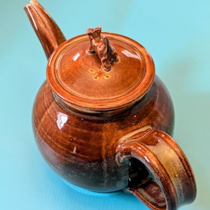 May include: A brown ceramic teapot with a lid and handle. The teapot has a glossy finish with a gradient of dark brown to lighter brown. The lid features a small decorative element.