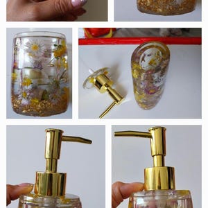 Peut inclure: Distributeur de savon transparent avec une pompe dorée, rempli de fleurs séchées et de paillettes dorées. Les fleurs comprennent des marguerites blanches et des fleurs jaunes. Le distributeur est rectangulaire avec des bords arrondis.