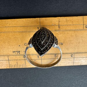 Op de afbeelding: Zilveren ring met een marquise-vormige voorkant, bezet met kleine, donkere edelstenen. De ring ligt op een houten liniaal met centimeter markeringen, die het ontwerp en de grootte van de ring benadrukken.