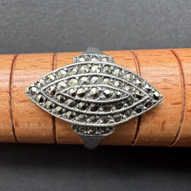 Op de afbeelding: Een zilverkleurige ring met een langwerpig ovaal ontwerp bezet met markasietstenen. Het middelpunt van de ring is een gedetailleerd patroon van kleine, donkere stenen. De ring wordt gepresenteerd op een houten oppervlak.