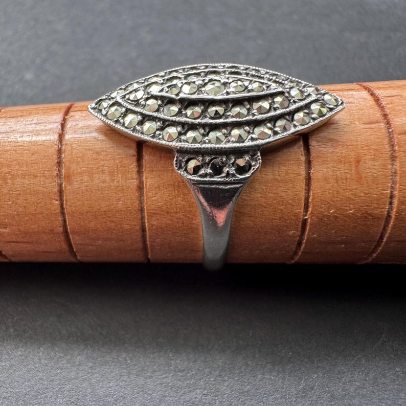 Op de afbeelding: Een zilverkleurige ring met een ovaal marcasietontwerp. De voorkant van de ring heeft meerdere rijen kleine, ronde marcasietstenen, wat een sprankelend effect cre&euml;ert. De band is van eenvoudig, gepolijst zilver. De ring wordt getoond op een houten deuvel.