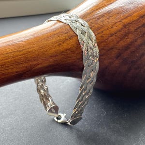 Könnte beinhalten: Ein silberfarbenes Armband mit einem mehrsträngigen geflochtenen Design und einer polierten, reflektierenden Oberfläche. Das Armband verfügt über einen sicheren Karabinerverschluss und ein kleines Metallschild in der Nähe des Verschlusses mit einer detaillierten Webstruktur.
