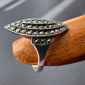 Op de afbeelding: Een vintage zilveren ring met een marquise-vormige voorkant. De ring is bezet met kleine, ronde, donkergekleurde stenen, gerangschikt in rijen. De band is van eenvoudig, gepolijst zilver. De ring wordt gepresenteerd op een houten oppervlak.