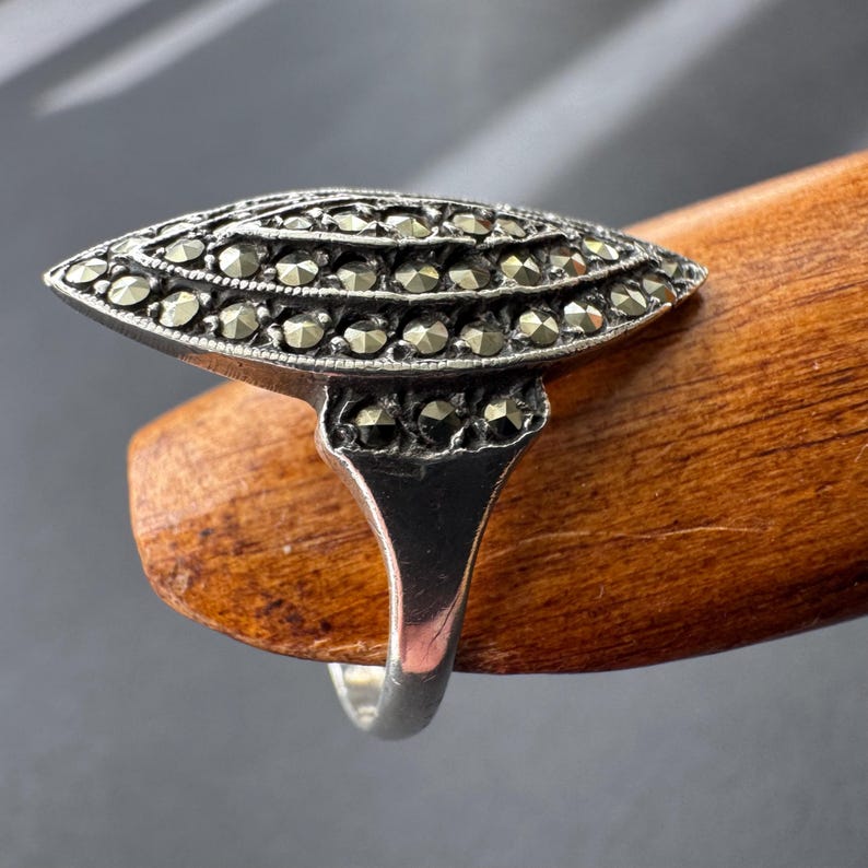 Op de afbeelding: Een vintage zilveren ring met een marquise-vormige voorkant. De ring is bezet met kleine, ronde, donkergekleurde stenen, gerangschikt in rijen. De band is van eenvoudig, gepolijst zilver. De ring wordt gepresenteerd op een houten oppervlak.