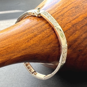 Può includere: Un braccialetto dorato con un design rettangolare. Il braccialetto presenta sezioni con piccoli abbellimenti scintillanti e una chiusura. Il braccialetto è esposto su una superficie di legno.