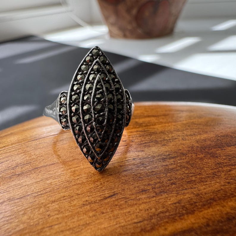 Op de afbeelding: Een zilverkleurige ring met een marquise-vormige voorkant, dicht bezet met kleine, donkere edelstenen. Het ontwerp van de ring heeft een centraal, langwerpig ovaal met symmetrische, gelaagde rijen stenen, geflankeerd door kleinere, afgeronde secties. De ring ligt op een houten oppervlak.