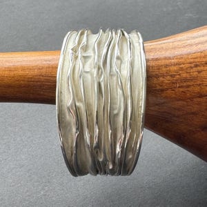 May include: A wide, silver ring with a textured, rippled surface. The ring is displayed on a wooden dowel against a grey background. The ring's design features vertical, wave-like patterns.