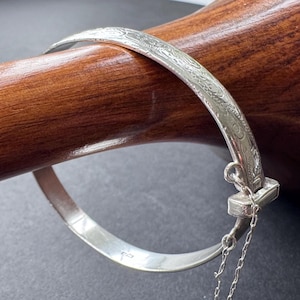 Könnte beinhalten: Ein silberner Armreif mit einem komplizierten, gravierten Blumenmuster auf der Außenfläche. Der Armreif verfügt über einen Klappverschluss mit einer zarten, befestigten Sicherheitskette, und ein kleiner Stempel ist auf dem Innenband sichtbar.