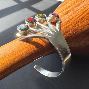 May include: A silver-toned cuff bracelet featuring a fan-like design with four prong-set faceted stones. The stones are arranged in a row, displaying alternating colors of dark green, deep red, olive green, and a final dark red gemstone at the end.