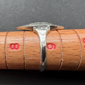 Op de afbeelding: Een zilveren ring met een Art Deco-ontwerp, met een centraal rechthoekig element met een reeks horizontale lijnen en kleine vierkante accenten. De ring wordt getoond op een houten ringmaat, die de maat en vorm laat zien.