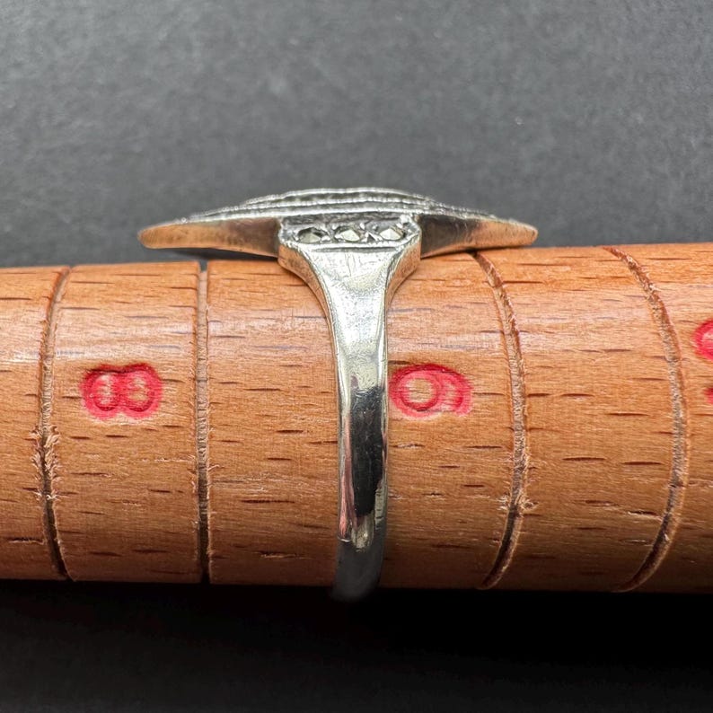 Op de afbeelding: Een zilveren ring met een Art Deco-ontwerp, met een centraal rechthoekig element met een reeks horizontale lijnen en kleine vierkante accenten. De ring wordt getoond op een houten ringmaat, die de maat en vorm laat zien.