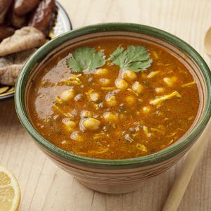 Peut inclure: Un bol de soupe aux pois chiches et aux herbes, servi avec une cuillère en bois et une assiette de dattes et de figues. La soupe est d'une riche couleur orangée et garnie d'herbes vertes fraîches. Une tranche de citron est visible.