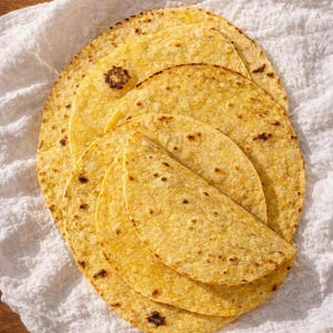 Könnte beinhalten: Ein Stapel runder, gelber Mais-Tortillas mit sichtbaren gerösteten braunen Flecken. Die Tortillas sind übereinander geschichtet, einige sind gefaltet, und zeigen eine texturierte Oberfläche sowie eine goldgelbe Farbe der runden Fladenbrote.