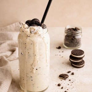 Peut inclure: Un grand verre rempli d'un milkshake blanc crémeux avec des miettes de biscuits noirs, surmonté de biscuits sandwich au chocolat entiers et d'une paille noire. Le milkshake déborde sur les côtés du verre, accompagné d'un petit pot de biscuits.