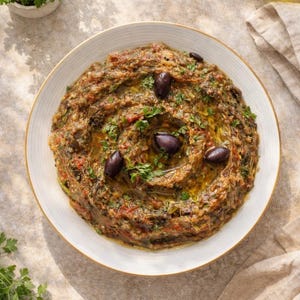 Peut inclure: Un bol blanc rond rempli d'une purée d'aubergines texturée, garnie d'olives violettes foncées et de persil vert frais. La préparation contient un mélange d'aubergines rôties, de morceaux de tomates rouges et d'herbes, arrosée d'huile d'olive.