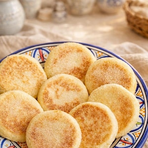 Peut inclure: Une assiette de pâtisseries rondes et dorées, peut-être des muffins anglais ou des crumpets, disposées sur une assiette décorative bleue et blanche. Les pâtisseries ont une surface légèrement texturée et sont saupoudrées d'un fin enrobage. L'assiette est posée sur un tissu de ton neutre.