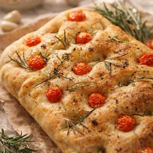 Peut inclure: Un pain focaccia rectangulaire doré, garni de tomates cerises rouge vif, de brins de romarin vert frais et d'herbes séchées. La surface du pain est alvéolée et légèrement huilée, ce qui lui donne un aspect rustique et appétissant.