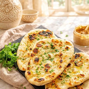 May include: Three pieces of golden-brown naan bread topped with melted butter and chopped green herbs. The flatbreads are arranged on a dark rectangular slate board, garnished with fresh parsley, and served alongside a small glass jar of dip.