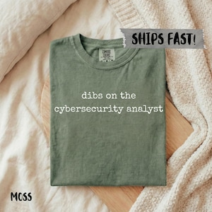 May include: A moss green t-shirt with the white text "dibs on the cybersecurity analyst". The t-shirt is folded on a wooden board. It has a crew neck and short sleeves.