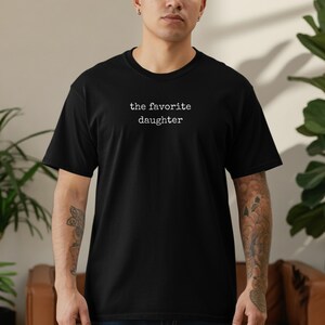 Black t-shirt with "the favorite daughter" text on chest, worn by a person.