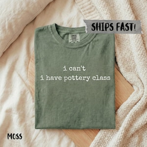 Puede incluir: Una camiseta verde musgo con el texto "i can't i have pottery class" en blanco. La camiseta está doblada sobre una tabla de madera, con una manta de tonos neutros en el fondo. La camiseta está etiquetada como "Comfort Colors".