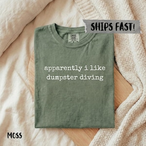 Op de afbeelding: Een mosgroen t-shirt met de tekst "apparently i like dumpster diving" in het wit. Het shirt is op een houten plank gevouwen, met een neutraal gekleurde deken op de achtergrond. Het shirt is gelabeld "Comfort Colors".