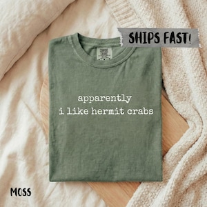 May include: A moss green t-shirt with the text "apparently i like hermit crabs" in white. The shirt is folded on a wooden cutting board, with a cream-colored blanket in the background. The shirt is labeled "Comfort Colors".