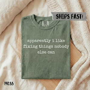 May include: A moss green t-shirt with the text "apparently i like fixing things nobody else can" in white. The shirt is folded on a wooden board, with a neutral-toned background.