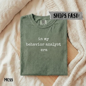 Op de afbeelding: Een mosgroen t-shirt met de witte tekst "in my behavior analyst era". Het shirt is op een houten plank gevouwen, met het woord "MOSS" linksonder. Het shirt is gemaakt van een zacht materiaal.