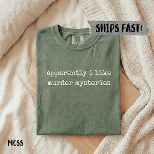 May include: Moss green t-shirt with the text "apparently i like murder mysteries" in white. The shirt is folded on a wooden board. The shirt is made of a soft material and has a crew neck.
