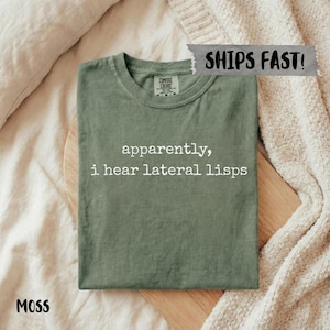 May include: A moss green short-sleeve t-shirt with a crew neckline. The shirt features the white printed text "apparently, i hear lateral lisps" centered on the chest. The garment includes a Comfort Colors brand label inside the neck collar.