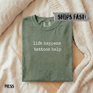 May include: A moss green t-shirt with the text "life happens tattoos help" in white. The shirt is folded on a wooden board, with a neutral-toned background. The shirt has a crew neck and short sleeves.