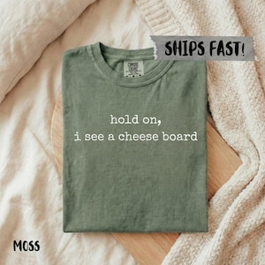 May include: A moss green t-shirt with the text "hold on, i see a cheese board" in white. The shirt is folded on a wooden cutting board. The shirt has a crew neck and short sleeves. The word "MOSS" is printed in the bottom left corner.