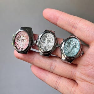 May include: Three silver-toned rings with oval faces, each featuring a floral design. The ring faces are in shades of pink, white, and blue. The rings are held in a hand.