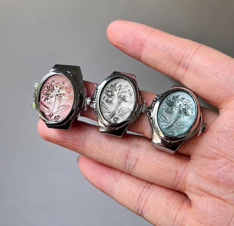 May include: Three silver-toned rings with oval faces, each featuring a floral design. The ring faces are in shades of pink, white, and blue. The rings are held in a hand.