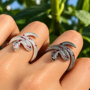 Puede incluir: Dos anillos con forma de serpiente, uno con acabado en plata brillante y otro en plata oxidada oscura. Ambos anillos presentan escamas texturizadas y pequeños detalles en esmalte rojo a lo largo del cuerpo, con las cabezas en la parte frontal.