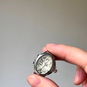 May include: A silver-toned ring with a clear, oval-shaped face. The face features a detailed, etched design of a tree with leaves and flowers. The ring has a decorative band and a small, adjustable mechanism.