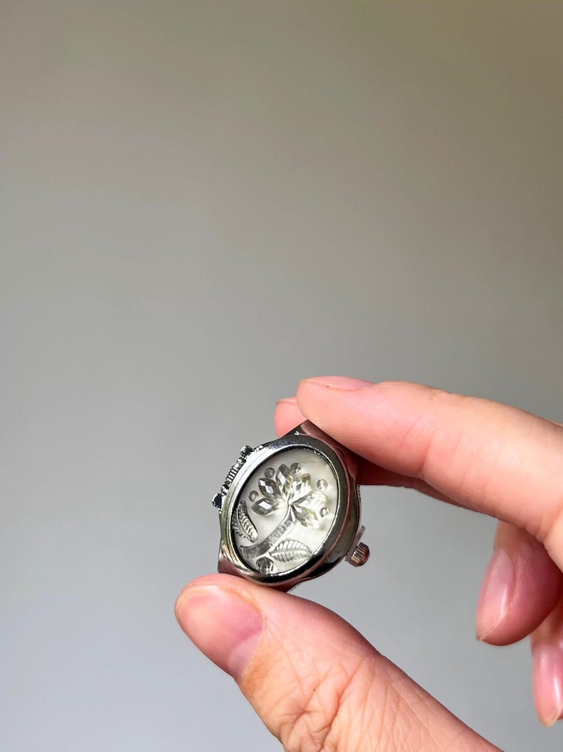 May include: A silver-toned ring with a clear, oval-shaped face. The face features a detailed, etched design of a tree with leaves and flowers. The ring has a decorative band and a small, adjustable mechanism.