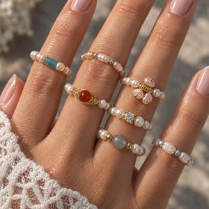 May include: A collection of six beaded rings featuring white freshwater pearls, small metallic beads, and various centerpieces including a blue rectangular bead, a red round stone in gold wire, a clear crystal, and a grey round stone.