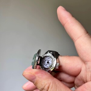 May include: A silver-toned ring watch with a hinged cover. The watch face is white with black numerals and hands. The ring has a decorative bezel and a black band. The hinged cover has a light blue stone.